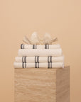 Kobn Crema Bathroom set includes towels, bathmat and handtowel, folded on a travertine plinth with a clam shell on top.