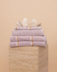 Folded stack of Lilac Kobn towels, hand towel and bathmat on a travertine plinth and topped with a clam shell.