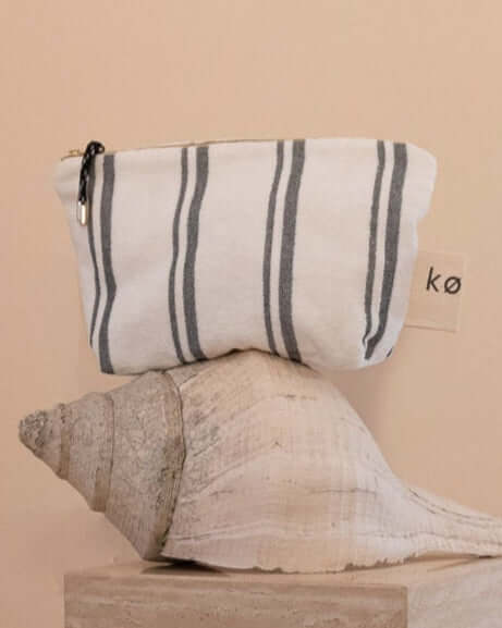 Kobn towelling Crema pouch perched on a large conch shell with a brand label, gold zip and tassle. For use as a clutch or organiser.