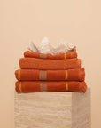 Stack of textured luxury towels in a Tamarillo colour featuring stripes and raw edge fringing on a travertine plinth with a clam shell on top