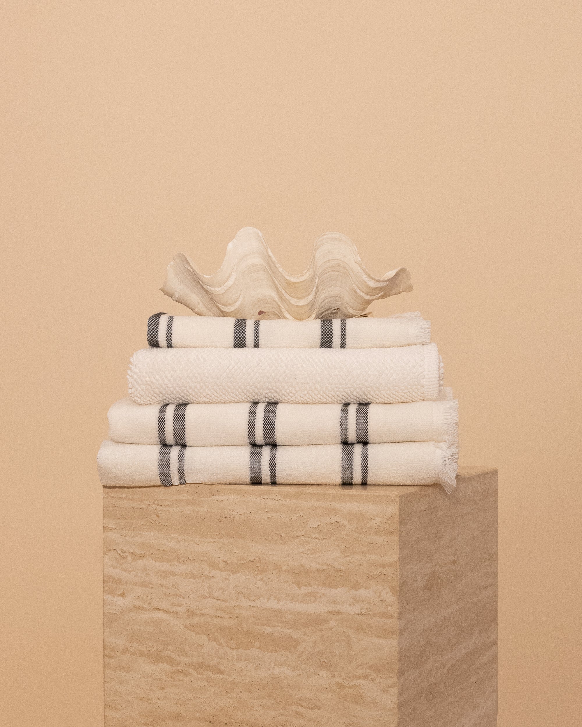Kobn Crema Bathroom set includes towels, bathmat and handtowel, folded on a travertine plinth with a clam shell on top. 