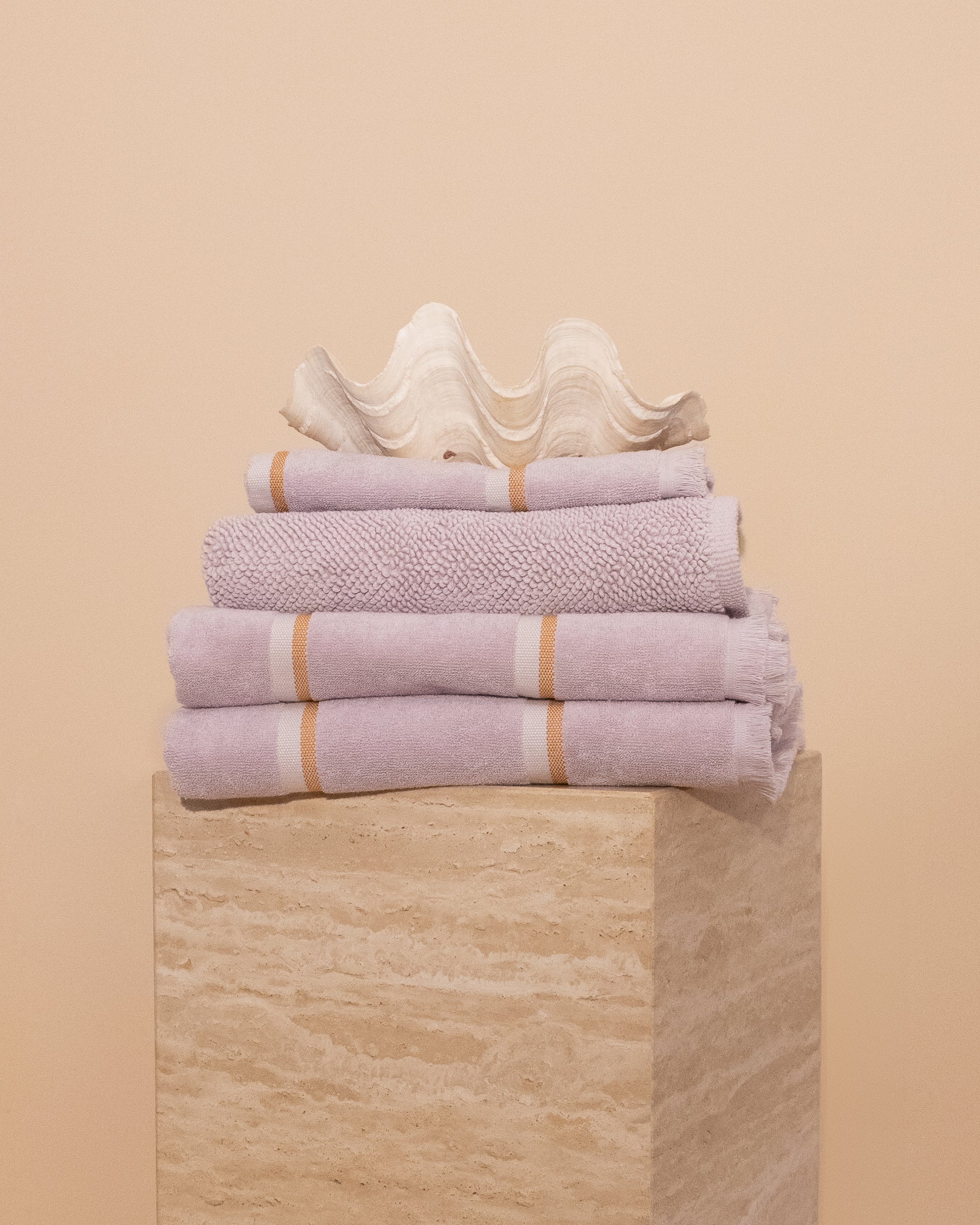 Folded stack of Lilac Kobn towels, hand towel and bathmat on a travertine plinth and topped with a clam shell. 