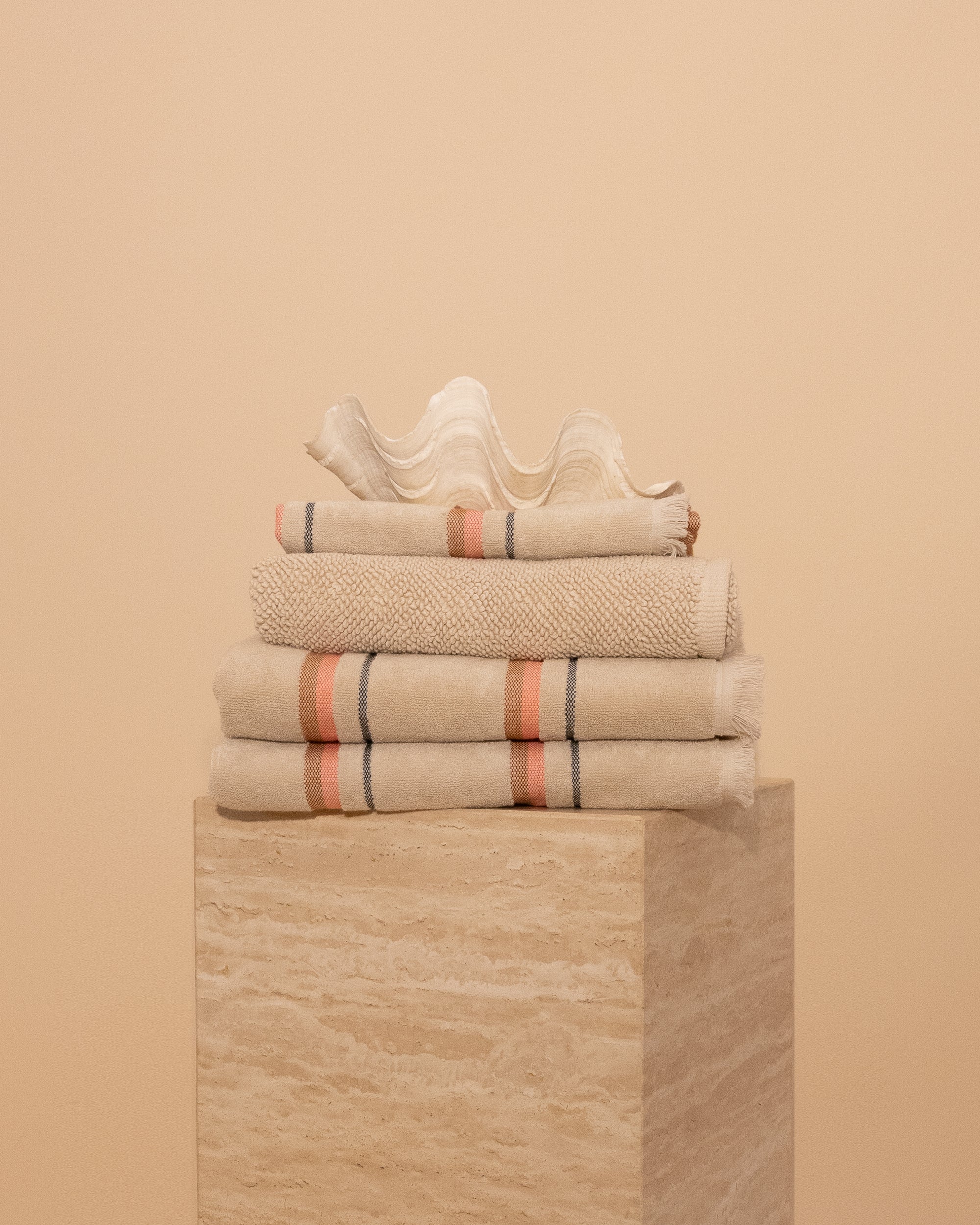 Folded Kobn Sand towels, bathmat and hand towel on a travertine plinth featuring the stripe and fringe details. 
