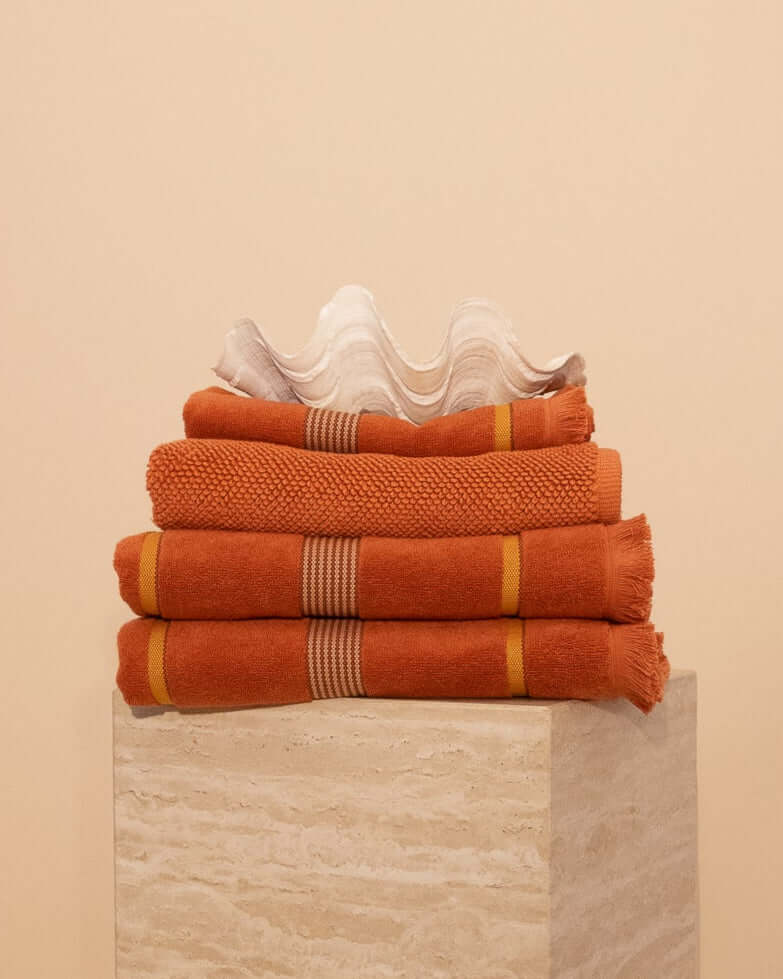 Stack of textured luxury towels in a Tamarillo colour featuring stripes and raw edge fringing on a travertine plinth with a clam shell on top