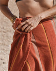 A woman with a black bikini top with a Tamarillo striped towel wrapped around her waist. 
