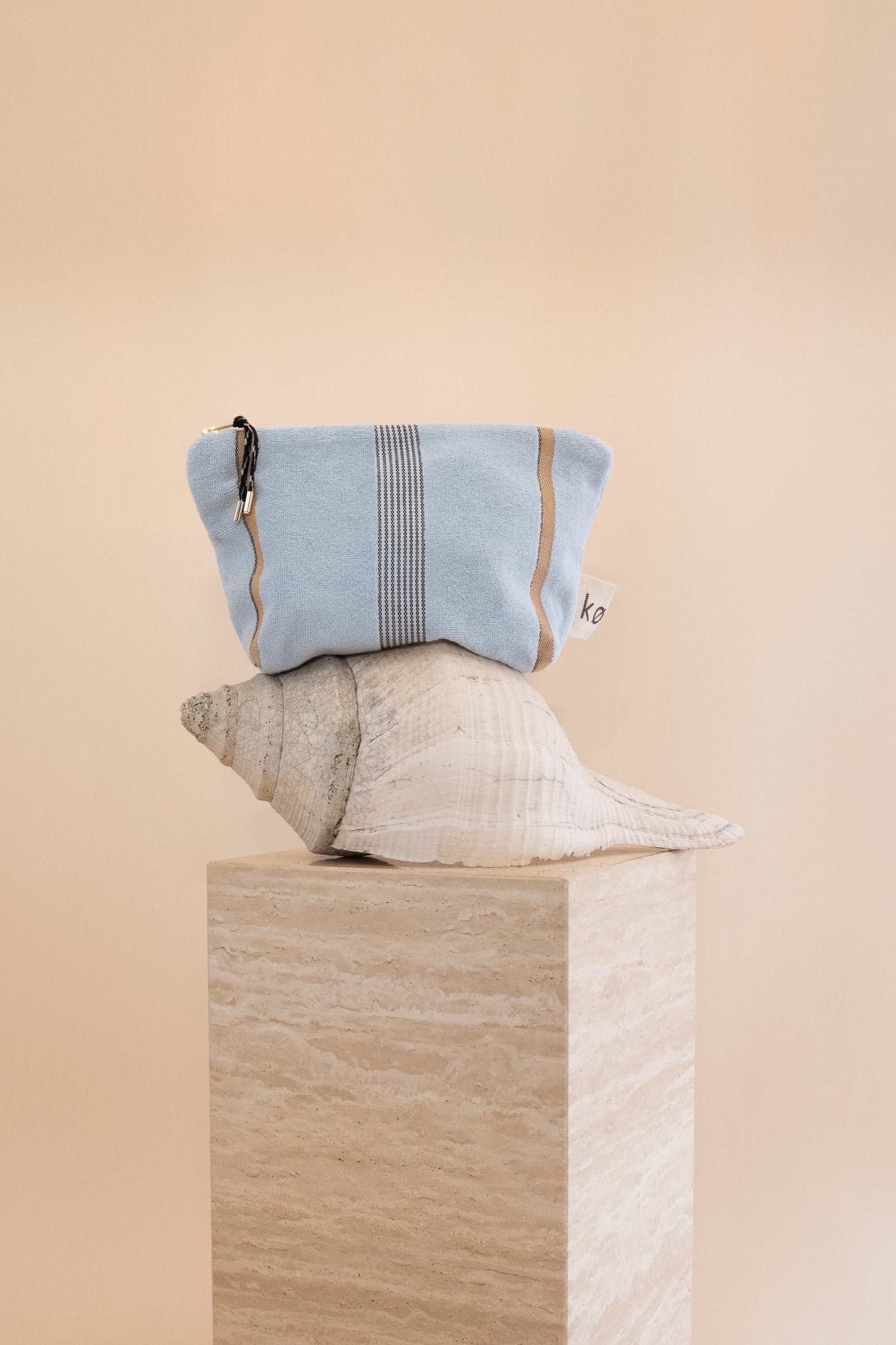 Light blue towelling pouch with stripes styled on a large shell and travertine plinth