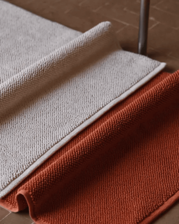 Two bathmats in Ecru and Tamarillo on floor with chunky cotton texture. 