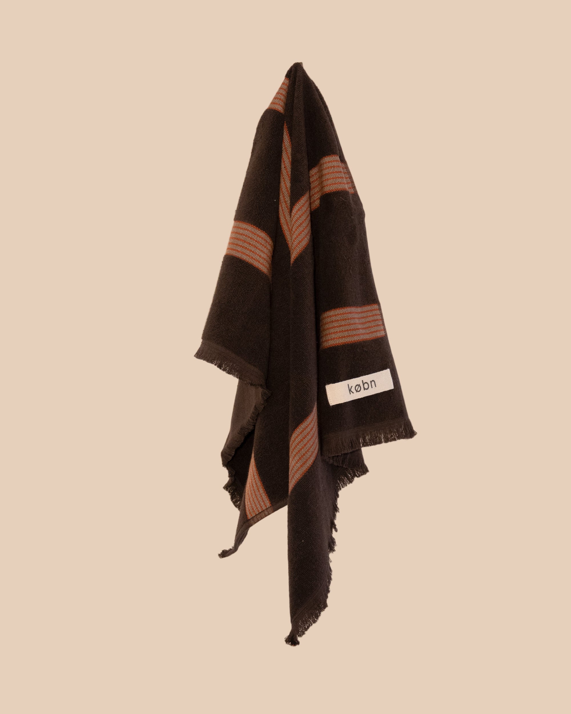 Chocolate brown hand towel with fringing and red and cream stripes hanging on a neutral background. 