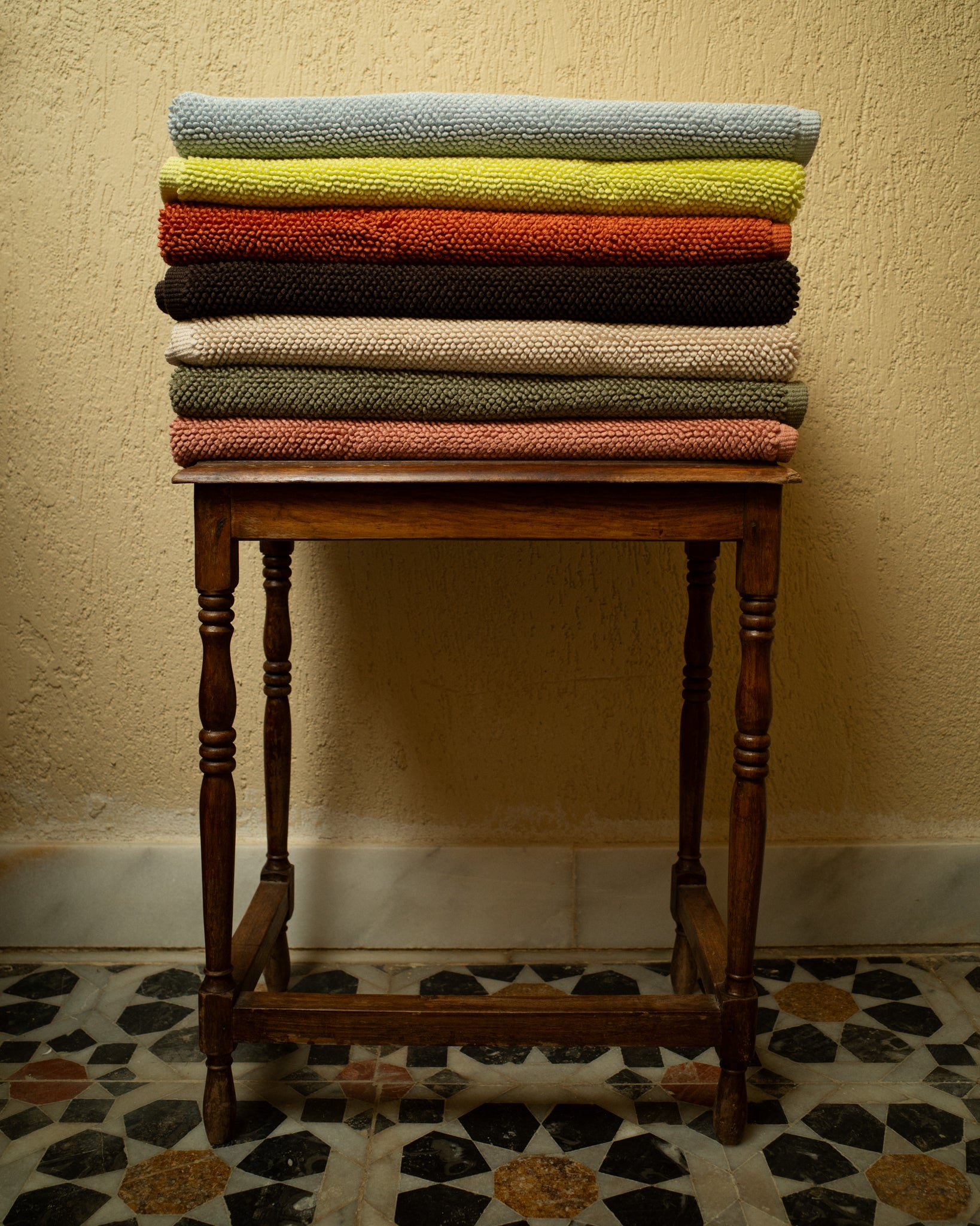 Stack of colorful folded bathmats on a wooden stool against a textured wall.