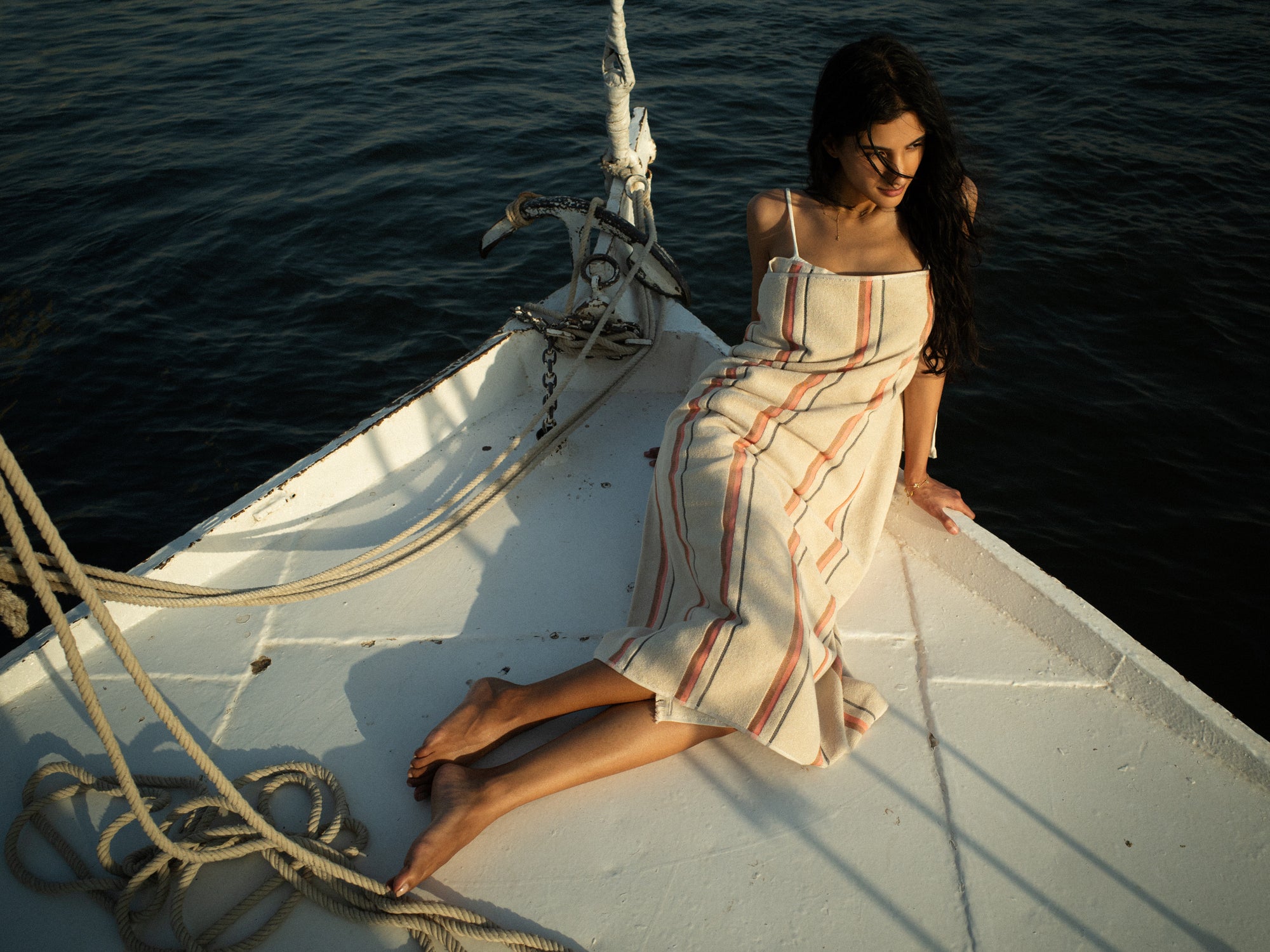 Woman wrapped  in a striped towel sitting on the front of a boat at sunset.