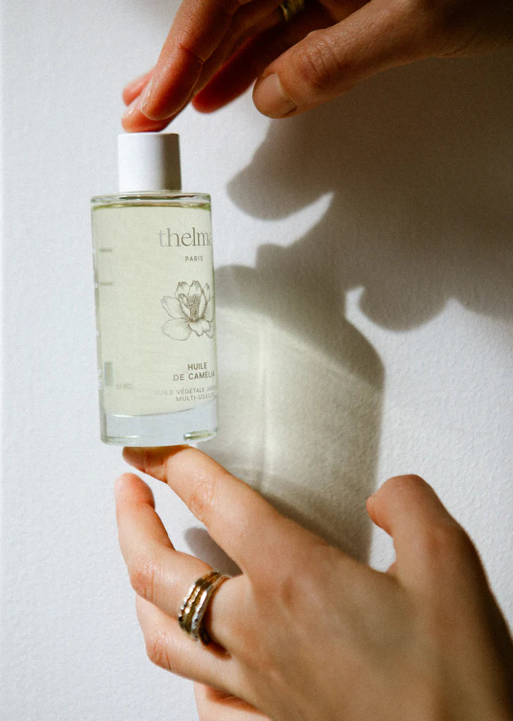Person holding a bottle of ThelmaCamellia oil against a white wall.